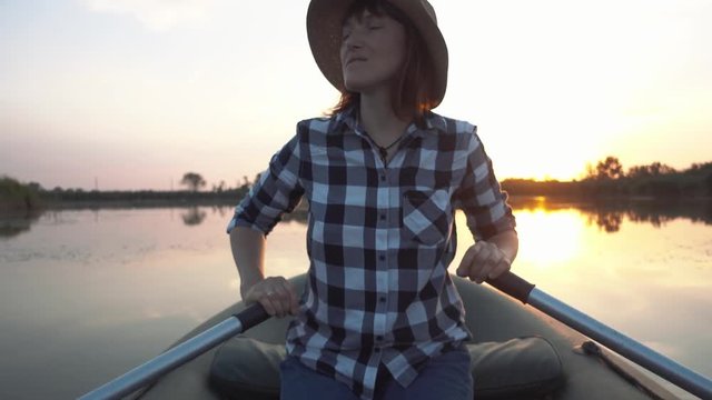 Young happy girl in a hat sails on an inflatable boat with wooden oars on the lake.