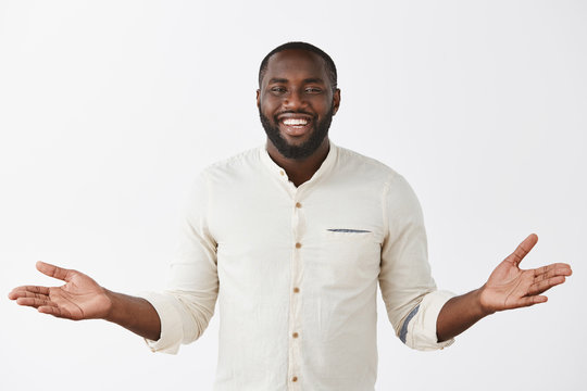 Indoor shot of charming carefree and happy cute dark-skinned bearded man in white shirt spreading palms aside and smiling broadly, inviting friend to come in, being hospitable and polite