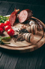 Smoked salami on a black table and a brown board. Sausages with rosemary, garlic and pepper and tomato cherry, dill, parsley.