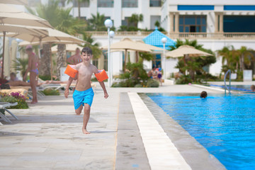 Smiling Caucasian boy in floating sleeves running along swimming pool  at resort.