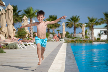 Joyful European boy running ang jumping along swimming pool at resort in summer. His arms are wide open.