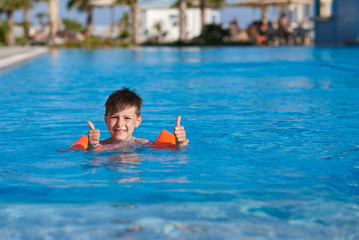 Smiling Caucasian boy in floating sleeves swimming in pool at resort. He is looking into camera and holding thumbs up.