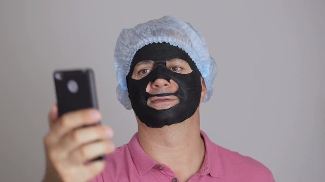 A man, applying a rejuvenating cosmetic mask of black cloth to his face and doing selfie with a smartphone