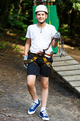 Happy, cute, young boy in white t shirt and helmet having fun and playing at the adventure park, holding ropes and climbing wooden stairs. Hobby, active lifestyle concept