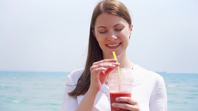 Young vegan woman drinking strawberry smoothie against sea in slow motion. Fit vegetarian female enjoying healthy lifestyle outdoors. Smiling girl doing thumbs-up gesture