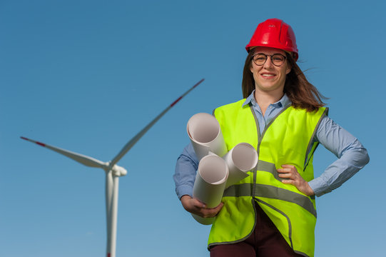 Positive young female engineer in red helmet and glasses with projects and plans against the background of a windmill and blue sky