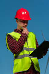 Serious young male chief engineer with a red helmet and green waistcoat signs the project agreement against a background of windmills and a blue sky