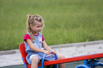 Small cute young blond child girl sits on see- saw swing on warm sunny day on bright green blurred...