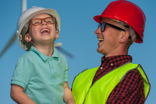 Engineer in the second generation. Portrait of a little positive boy in the hands of his father's engineer's in glasses and helmets against the background of windmill and blue sky