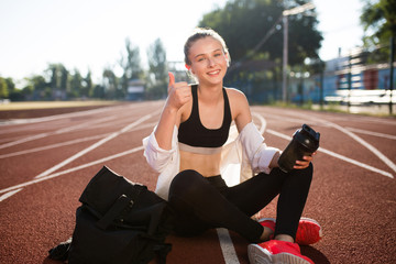 Smiling girl in wireless earphones holding sport bottle in hand joyfully looking in camera while showing thumb up on running track of stadium