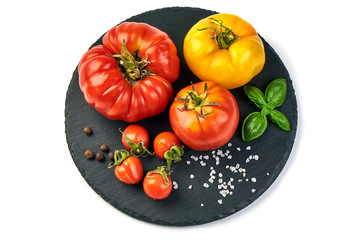 Colorful yellow and red tomatoes of different sizes and kinds, on rustic slate stone plate, with spices an basil leaves, isolated on white background. Top view.