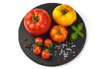 Colorful yellow and red tomatoes of different sizes and kinds, on rustic slate stone plate, with spices an basil leaves, top view, isolated on white background.