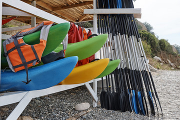 Kayaks, life jackets, oars under a wooden roof on the beach.