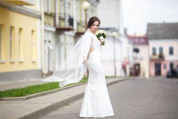 Fototapeta na wymiar young beautiful bride girl with wedding bouquet of roses stands on the street. the wind blows the veil