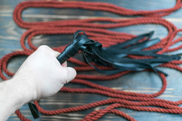 Scourge for BDSM in a man's hand and a rope for tying on a dark background. Accesories for sexual...