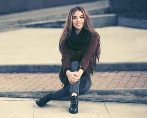 Happy young fashion woman in leather jacket sitting on sidewalk in city street
