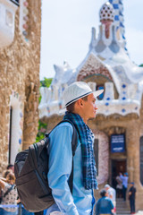 A young guy stands in a white hat and scarf with backpack near a gingerbread house in Guell Park, inside