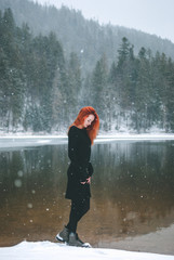 Handsome redhead girl looking down. Snowflakes falling on her hair. Splendid shady black mountain lake. Dreamy winter landscape of Carpathian mountains.
