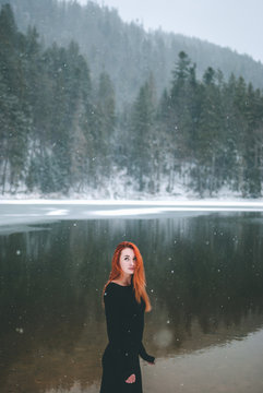 Handsome redhead girl looking up on the snowflakes falling on her hair. Splendid shady black mountain lake. Dreamy winter landscape of Carpathian mountains.