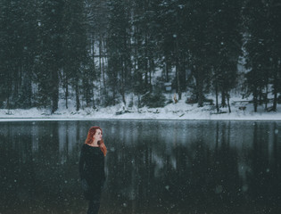 Fototapeta na wymiar Snowflakes flying over handsome black-eyed redhead girl looking in the distance. White snowflakes flying all around. Splendid shady black mountain lake