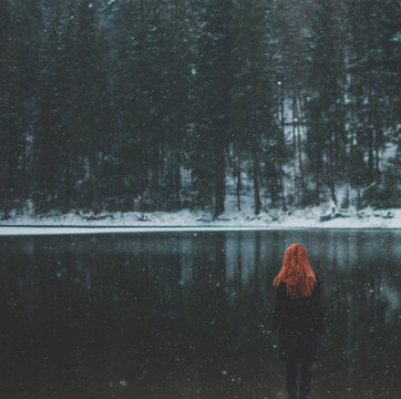 Square-shaped. Snowflakes flying over handsome black-eyed redhead girl looking in the distance. White snowflakes flying all around. Splendid shady black mountain lake. Look from behind
