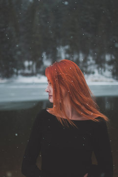 Handsome redhead girl looking in the distance. Snowflakes falling on her hair. Splendid shady black mountain lake. Dreamy winter landscape of Carpathian mountains.