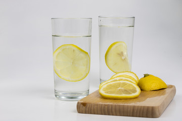 A glass of cold water with lemon on a white kitchen table. A refreshing drink in warm weather.