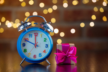 handmade gift box with purple bow and alarm clock on wooden table with fairy lights on bokeh background