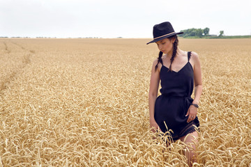 Naklejka na ściany i meble woman in a black dress and a hat stands in a yellow field with wheat in the summer