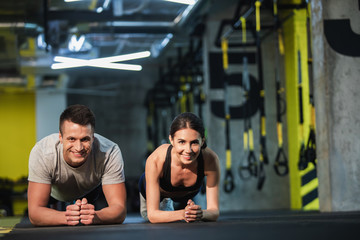 Merry man and woman are doing plank simultaneously in sport studio. They are balancing on elbows and looking at camera. Male and female are straining all body while exercising core