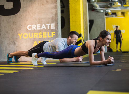 Cheerful man and woman are having joint workout in sport studio. They are planking on elbows synchronously for achieving better results. Motivation while exercising in sport class concept