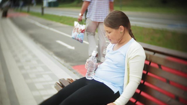 A small fat girl on rollers drinks water. A child drinks water in a park on a bench