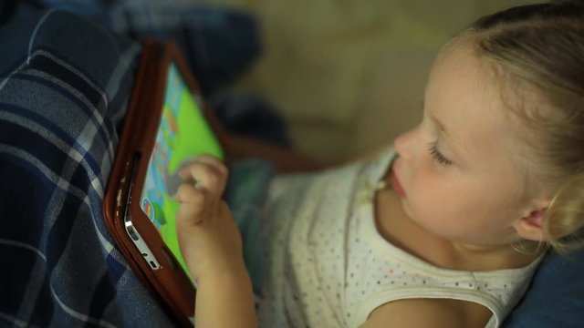 a little girl lying on a pillow in bed and playing games on a tablet.