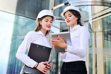 Two young cute girls in business clothes and white construction helmets discuss a business plan or...