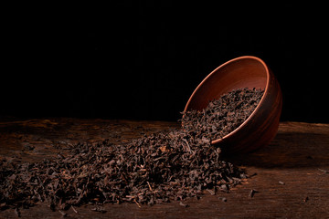 Dried black tea in clay bowl with sticks of cinnamon and a tea strainer isolated on black background