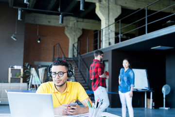Portrait of  creative young Middle-Eastern man wearing headphones and bright yellow shirt using laptop and listening to music while working in open space office of IT developers team, copy space