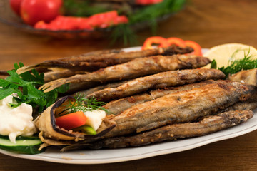 A small sea fish, fried entirely in oil, stacked on top of each other on a porcelain plate with stuffed eggplant, sliced fresh vegetables, decorated with sour cream and parsley.