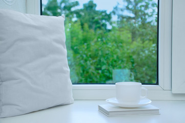 A white pillow, a book and a cup and saucer stand on the windowsill at the window in the summer.