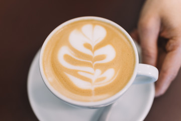 Latte art tree or heart pattern coffee in white mug with female hand holding on vintage brown table background, best aroma quality in the wake up morning