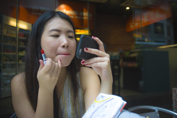 young happy and attractive Asian Korean tourist woman sitting at city coffee shop using lipstick makeup and mobile phone mirror waiting for online date