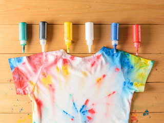 Tie dye style t-shirt and six cans of fabric paint on wooden table.