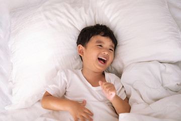 3 years old little cute Asian boy at home on the bed, kid lying playing and smiling on white bed with pillow and blanket, top view with copy space.