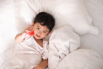 3 years old little cute Asian boy at home on the bed, kid lying playing and smiling on white bed with pillow and blanket, top view with copy space.