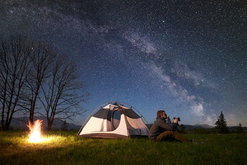 Incredible beautiful night camping in mountains. Young man hiker with photo camera sitting alone in...