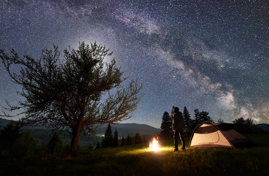 Camping site in mountain valley. Young hiker standing in front of tourist tent at burning campfire near big tree under night blue starry sky with Milky way and moon. Beauty of nature, tourism concept
