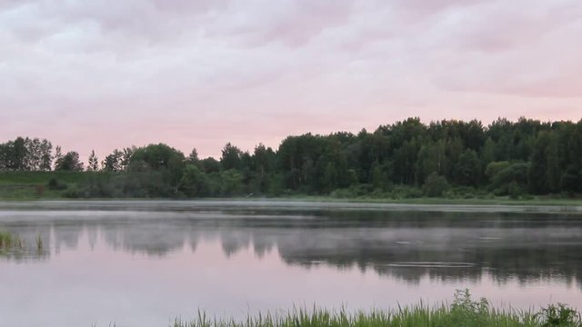 Beautiful dawn on the river, the fog moves over the water - summer view from the shore to the pond with trees on the far Bank