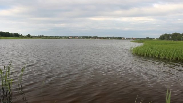 Light wind on the river, small waves on the water - summer view from the grassy shore to the pond with the village on the far shore
