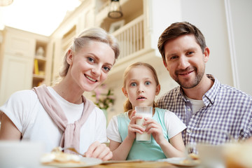 Shocked cute daughter with open mouth sitting among parents and holding full glass of milk, happy family members looking at camera in kitchen