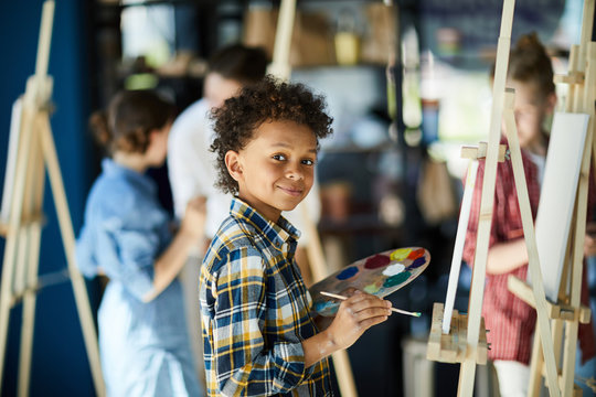 Young mixed-race boy with color palette standing by easel
