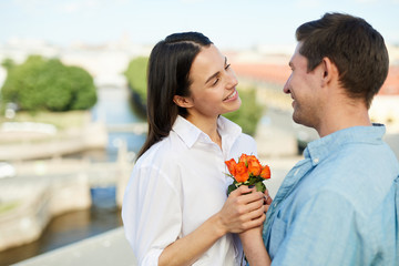 Happy young man in shirt giving pretty flowers to smiling beautiful girlfriend while they standing on roof and looking into eyes